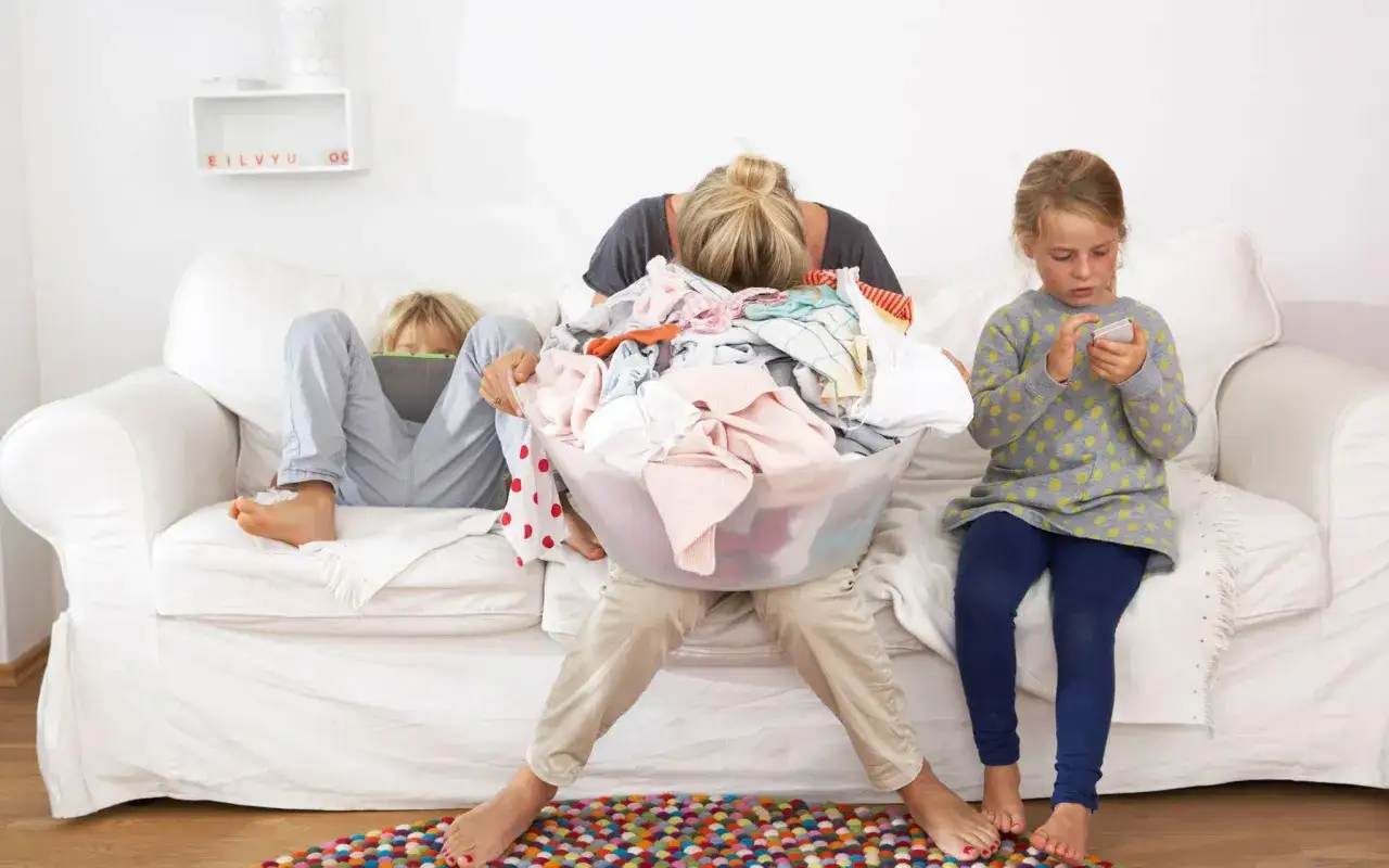 &Uuml;berforderte Mutter mit W&auml;schekorb, Kinder auf Sofa mit Tablets. Der **mental load deutsch** ist sp&uuml;rbar.