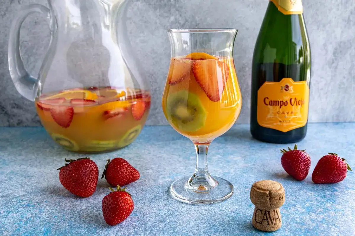 En glas cava sangria med frukt som jordgubbar och kiwi. En karaff med samma dryck och en flaska cava st&aring;r bredvid.