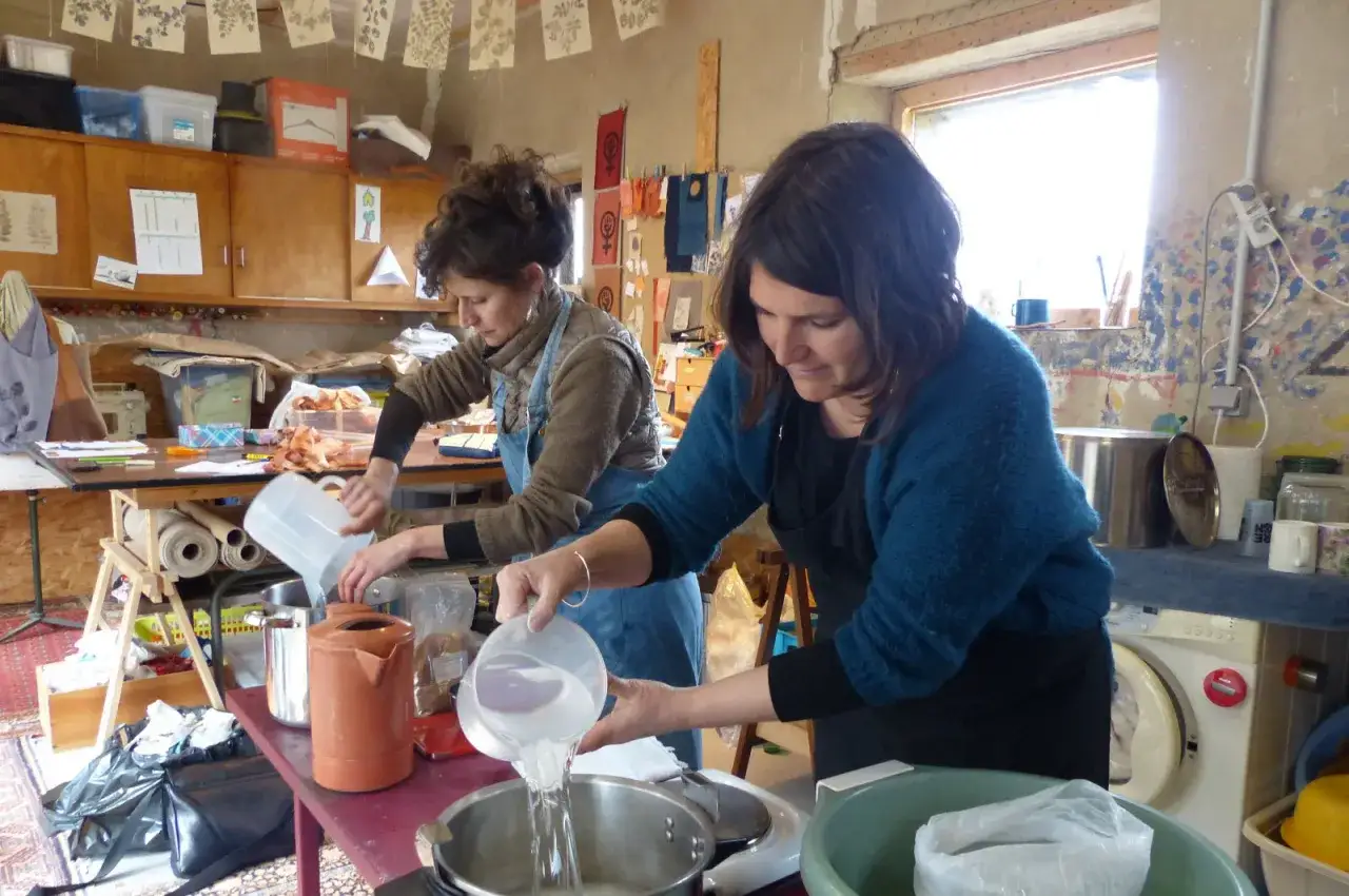 Deux femmes pr&eacute;parent la teinture dans un atelier. L'une verse de l'eau dans une marmite, l'autre ajoute des ingr&eacute;dients.