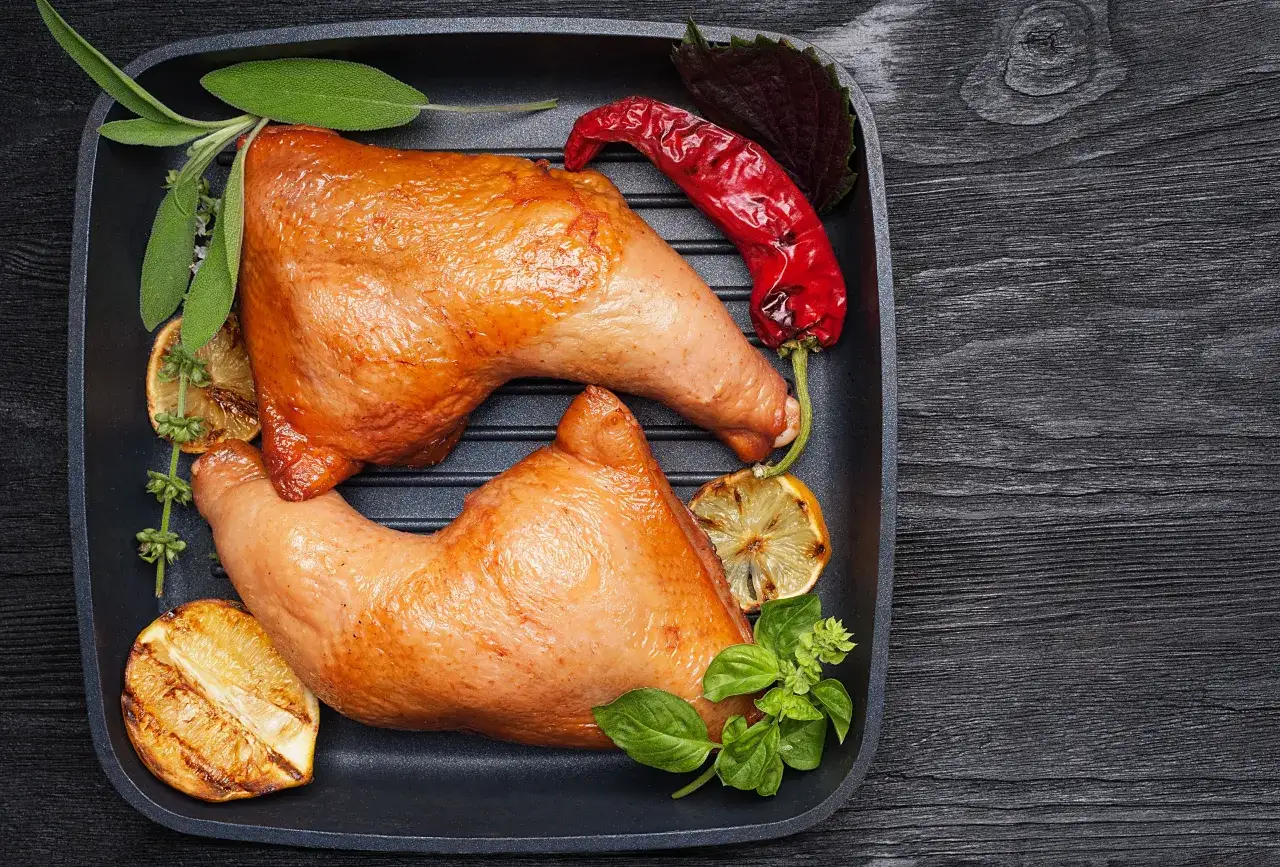 Dwa soczyste udka z kurczaka, wędzenie udek z kurczaka bez parzenia, podane z grillowaną cytryną, chili i ziołami na czarnej patelni grillowej.