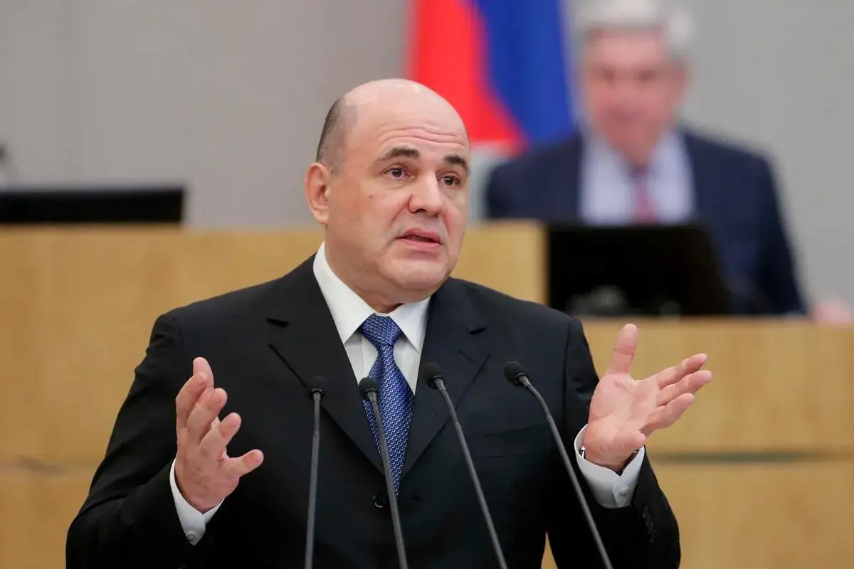 Premier Rosji, w czarnym garniturze i niebieskim krawacie, przemawia z podniesionymi dłońmi.