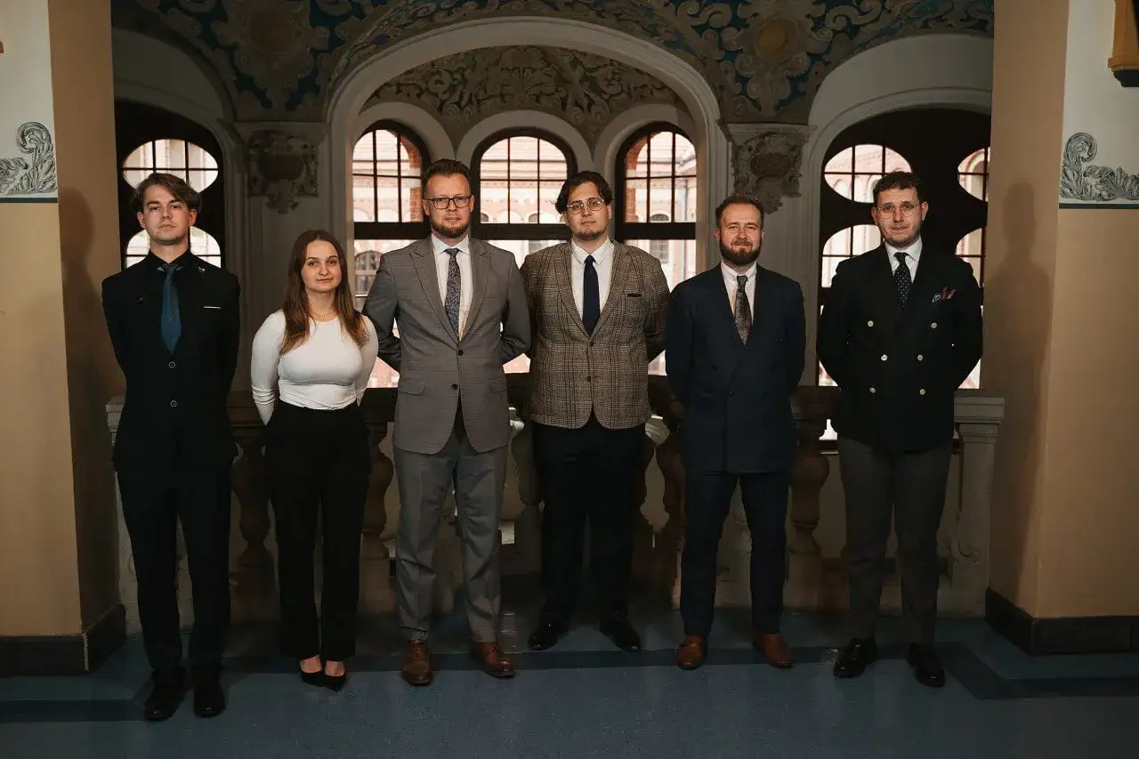 Sześć osób w eleganckich strojach stoi w rzędzie na tle zabytkowej architektury.