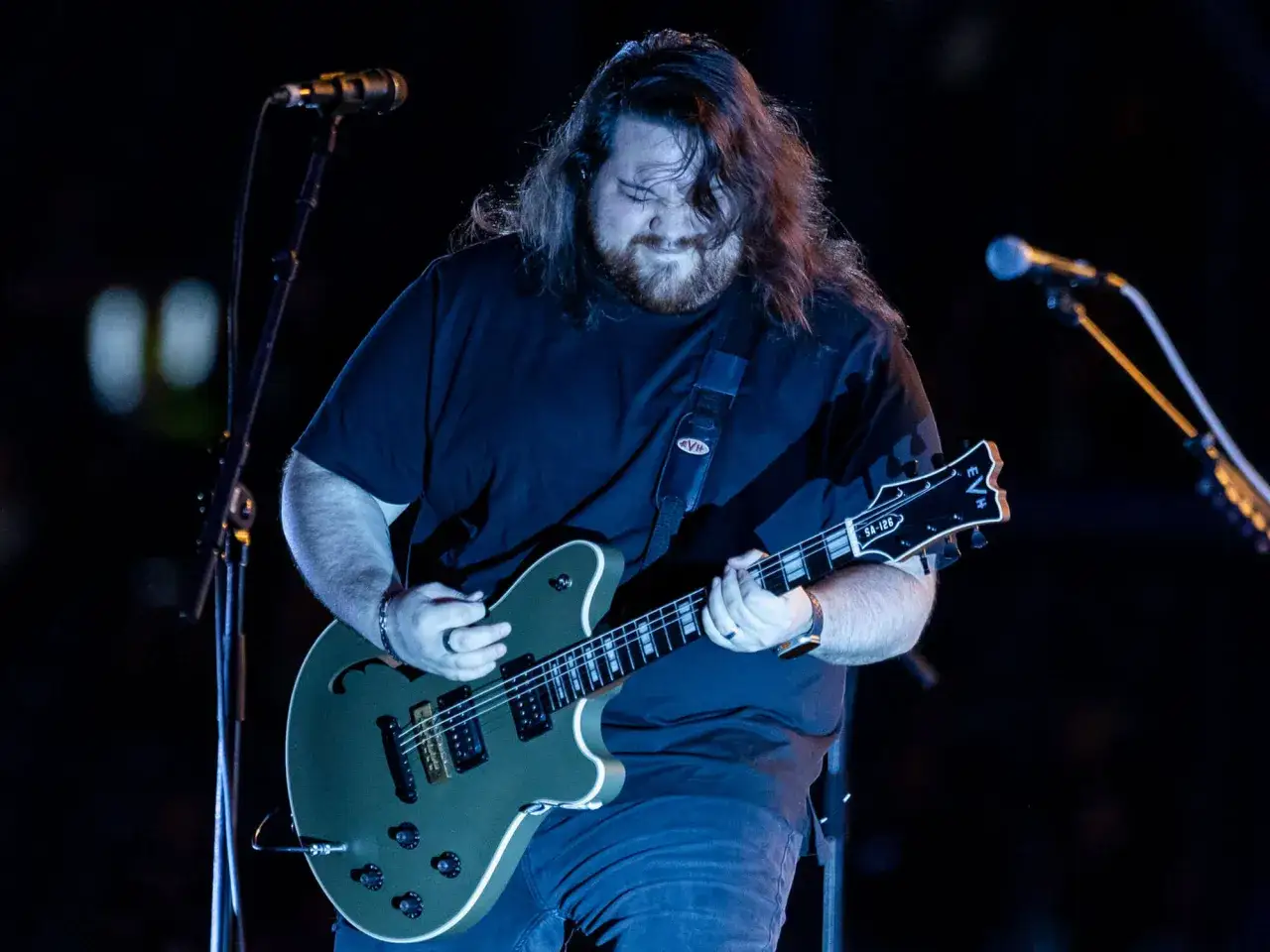Wolfgang Van Halen gra na gitarze na scenie, z długimi włosami i brodą, w czarnej koszulce.