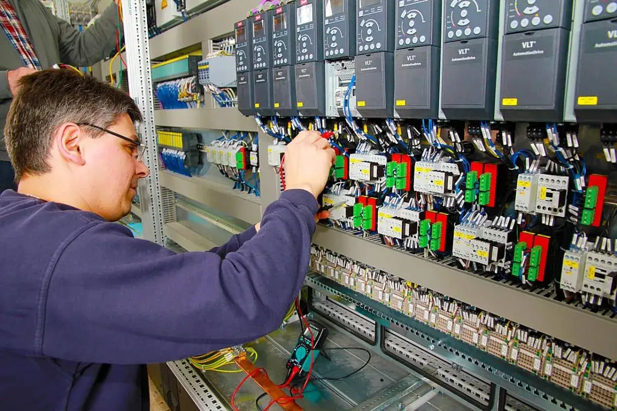 Elektryk pracuje przy panelu sterowania, podłączając przewody. Widać tu wiele przekaźników, falowników i elementów automatyki.