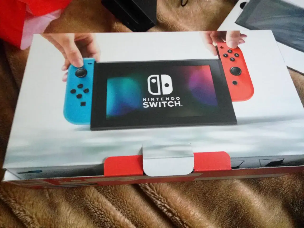 Dwie ręce trzymają niebieskiego i czerwonego Joy-Cona obok ekranu konsoli Nintendo Switch. Na pudełkach Nintendo Switch widać ten sam motyw.
