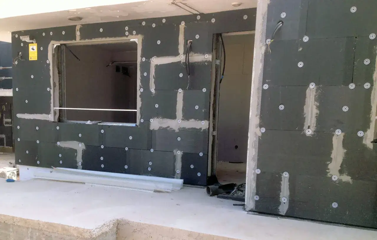Paneles de aislante térmico exterior fijados a una pared con mortero y fijaciones. Se aprecian un hueco para ventana y una entrada.