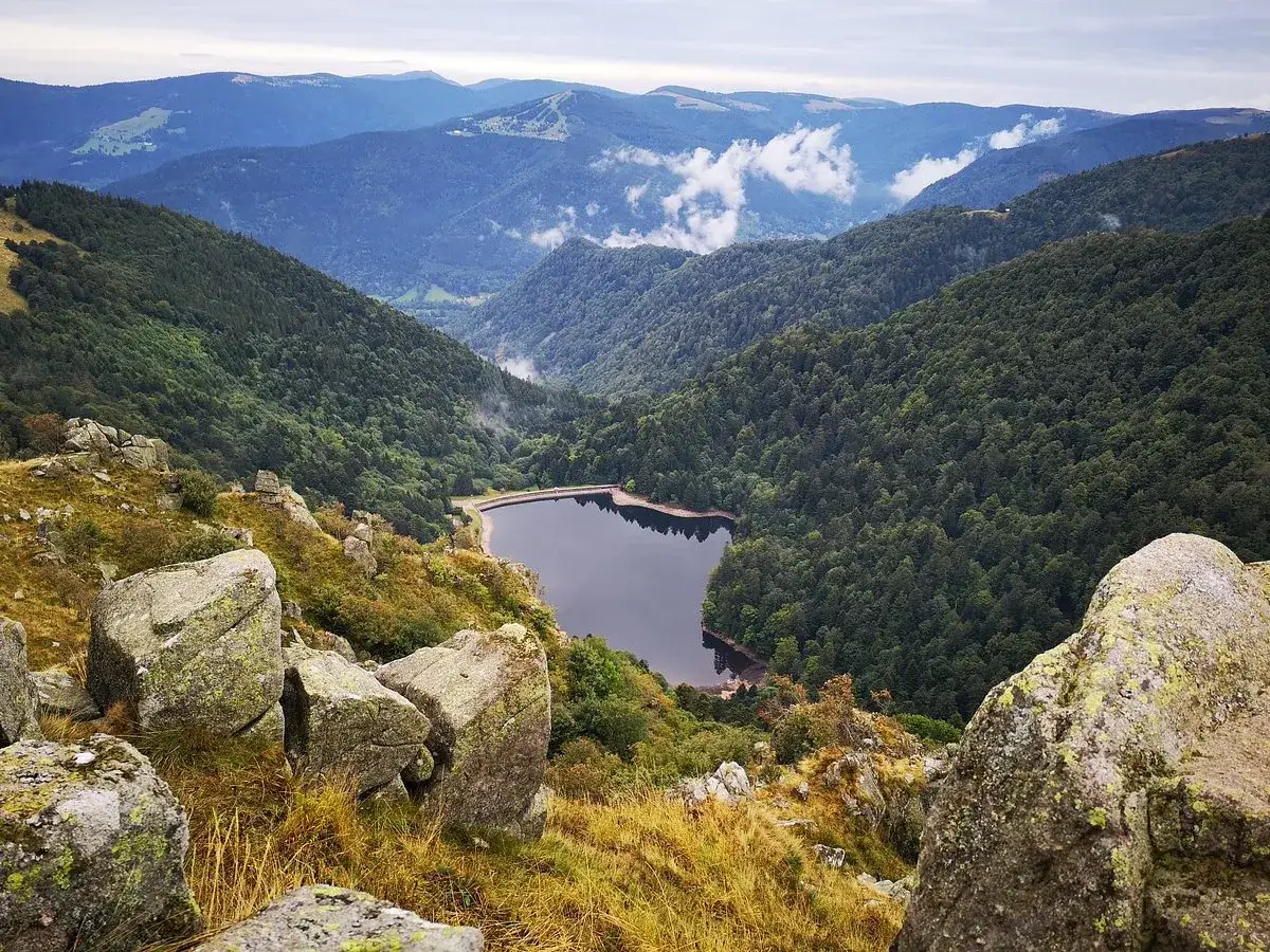 Vista panoramica dalla route de cretes su un lago incastonato tra montagne boscose. Rocce muschiate in primo piano.