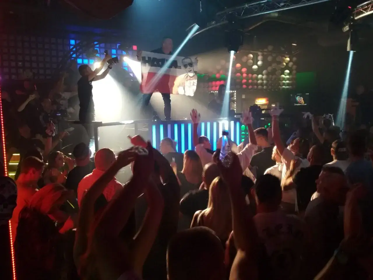 Tłum na koncercie DJ Hazel. Ludzie tańczą, jeden z nich trzyma flagę z podobizną DJ-a.