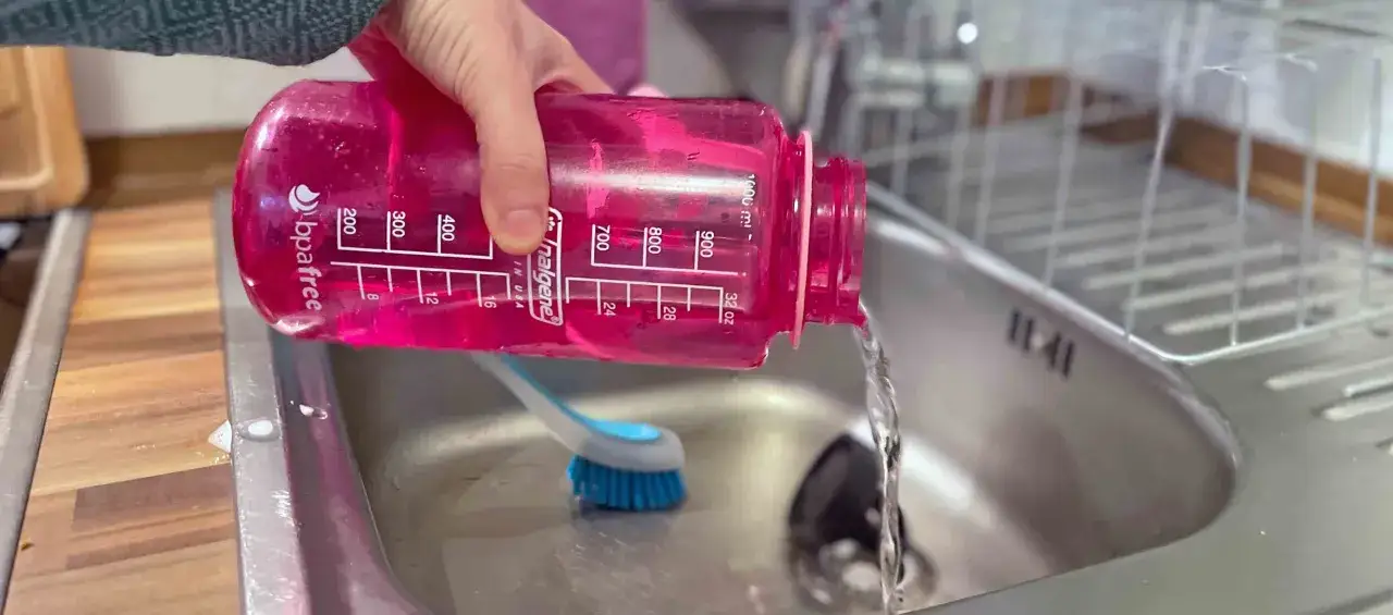 Eine Hand h&auml;lt eine pinke Trinkflasche &uuml;ber einem Sp&uuml;lbecken, w&auml;hrend Wasser herausl&auml;uft. Eine B&uuml;rste liegt daneben, bereit, die Trinkflasche zu reinigen.