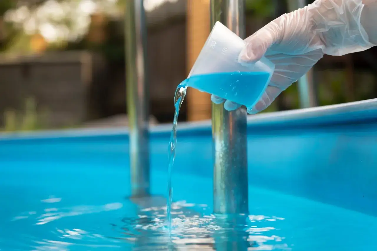 Gloved hand pours blue liquid from a measuring cup into a pool, a step in how to get crystal clear pool water.