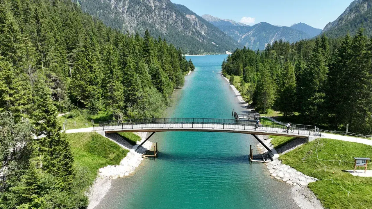 Radfahrer überqueren eine Brücke am türkisfarbenen Plansee, umgeben von grünen Wäldern und Bergen. Perfekt für Plansee wandern und Radtouren.
