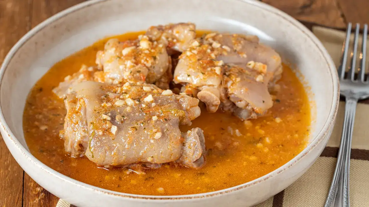 Manitas de cerdo en un cuenco de cer&aacute;mica, cubiertas con una sabrosa salsa de tomate y ajo. Un tenedor plateado descansa a la derecha.