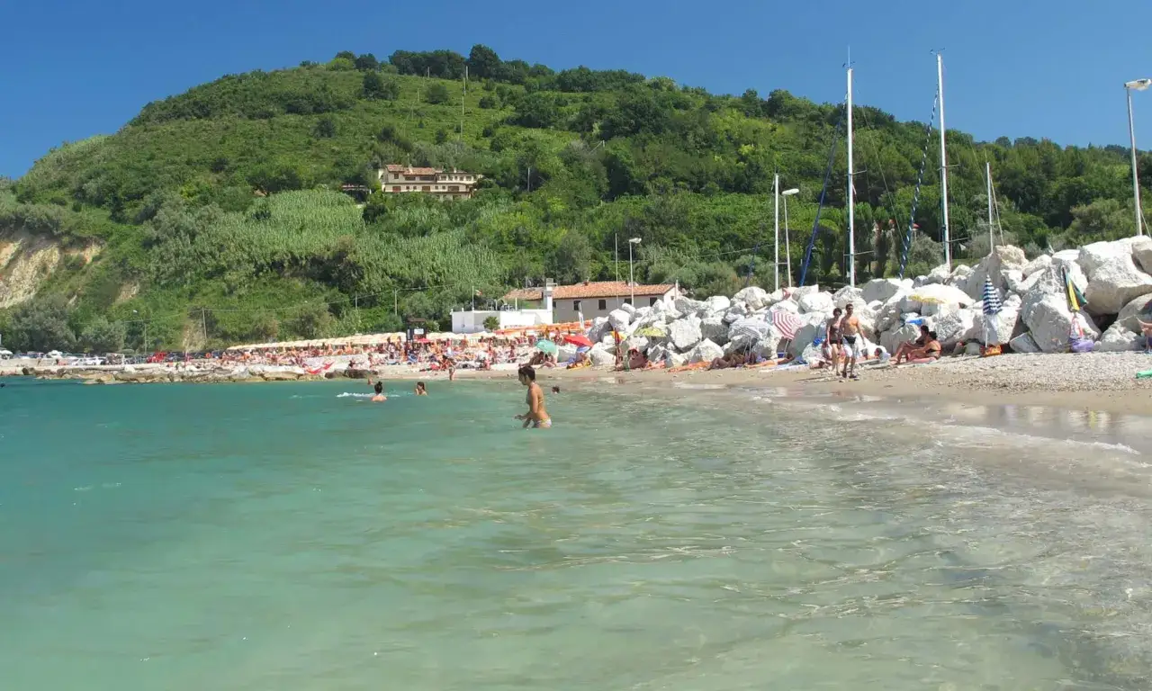 Spiaggia affollata a Gabicce Mare, con persone che nuotano e prendono il sole, colline verdi sullo sfondo.