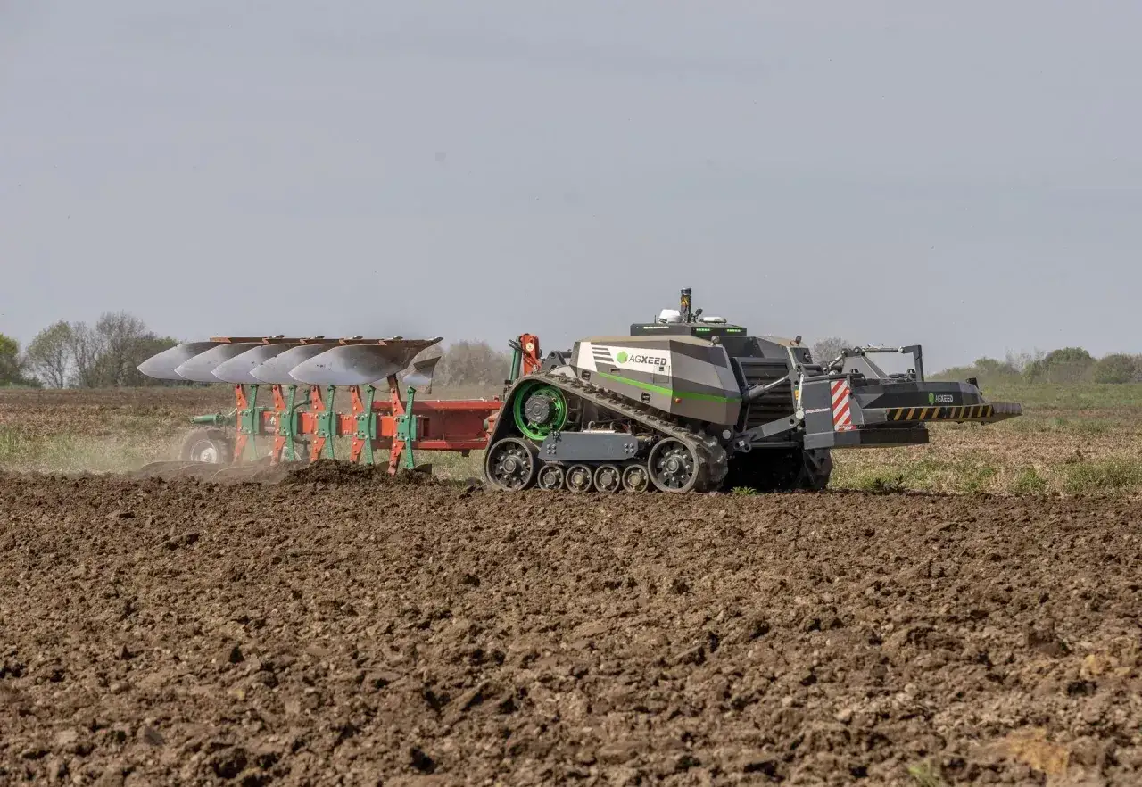 Autonomiczny traktor z gąsienicami i wieloskibowym pługiem do orania pracuje na polu, przygotowując ziemię.