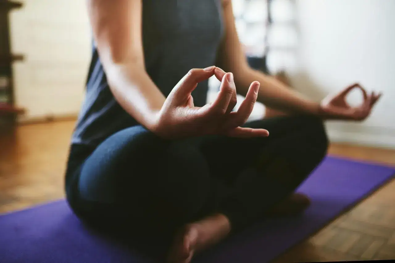 En person sitter i meditationsställning på en lila matta. Händerna vilar i knät med tumme och pekfinger sammanfogade, en vanlig gest för att visa hur man mediterar.