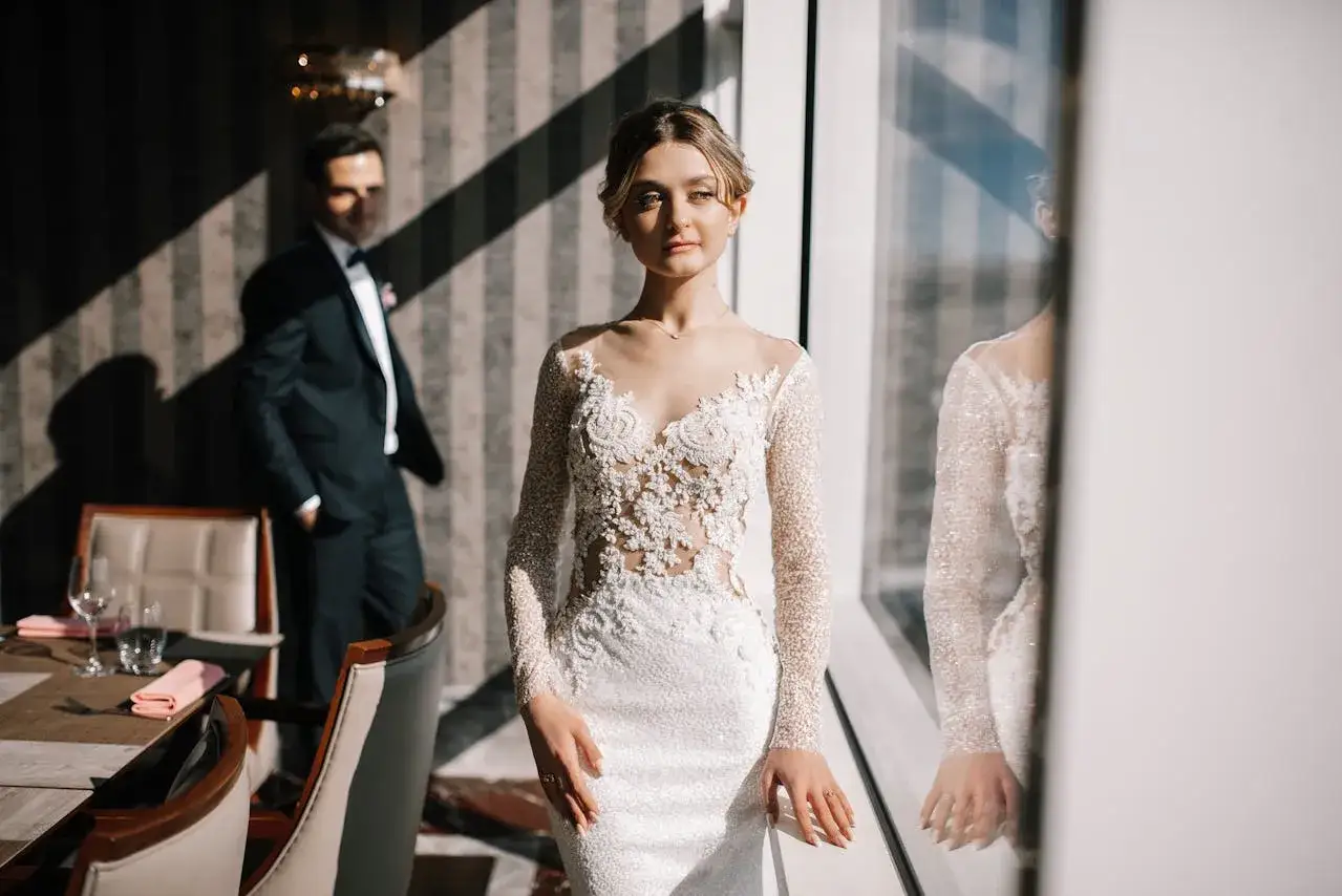 Elegancka panna młoda w minimalistycznej sukni ślubnej z koronkową górą i długimi rękawami. Obok stoi pan młody w garniturze.