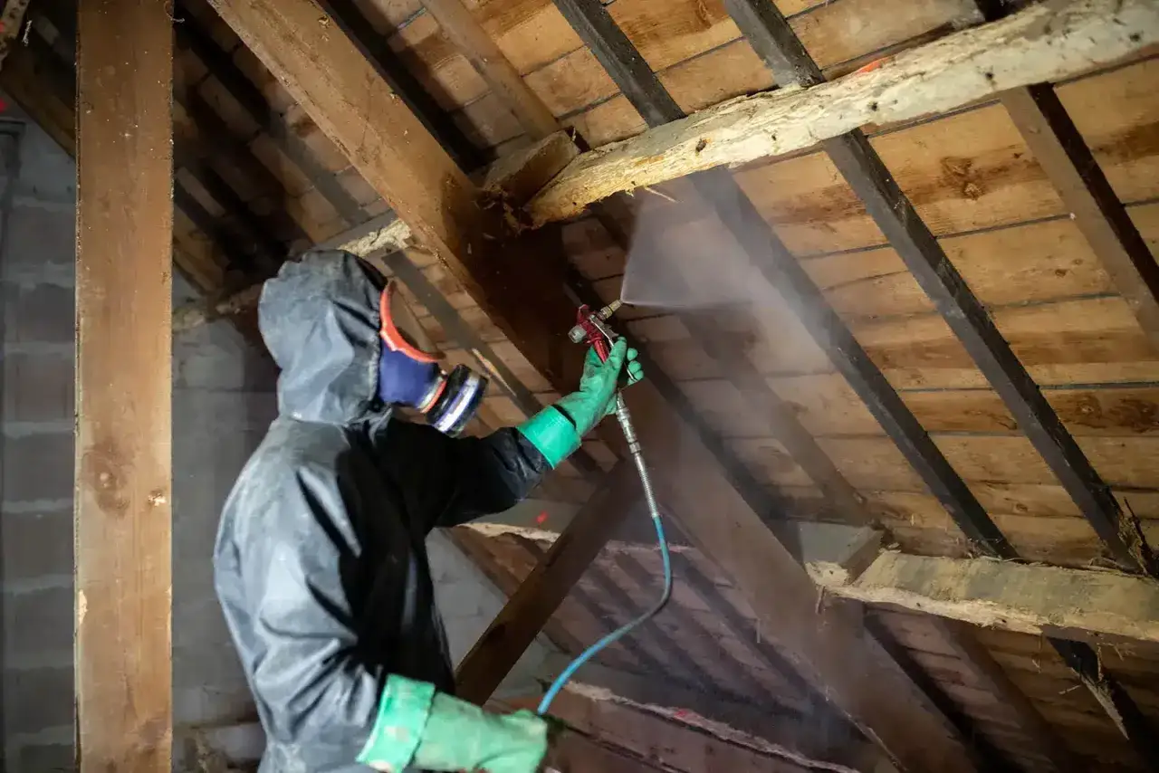 Person en combinaison et masque pulv&eacute;rise un produit sur la charpente en bois, &eacute;tape essentielle pour traiter charpente avant isolation.