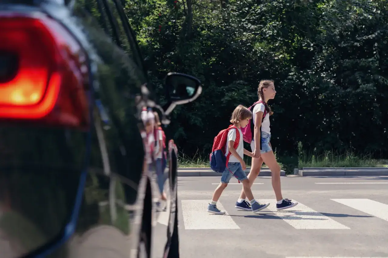 Bambini con zaini attraversano sulle strisce pedonali, rispettando le regole per i pedoni. Un'auto nera è parcheggiata a sinistra.