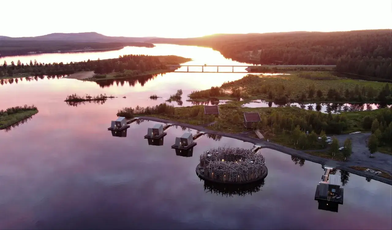 Unik arkitektur vid en sjö i Harads, Sverige. Flytande stugor och ett fågelbo-liknande hotell i solnedgången.