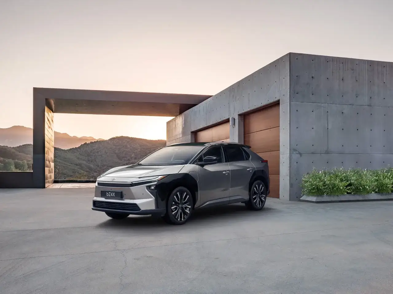 Nowoczesny Toyota bZ4X przed minimalistycznym domem z betonową elewacją i drewnianymi drzwiami garażowymi. Sprawdź jego cenę!
