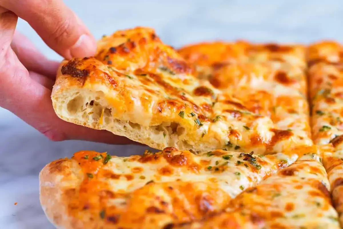Une main soulève une part de pinsa recette, garnie de fromage fondant et d'herbes. La pâte est aérée et dorée.