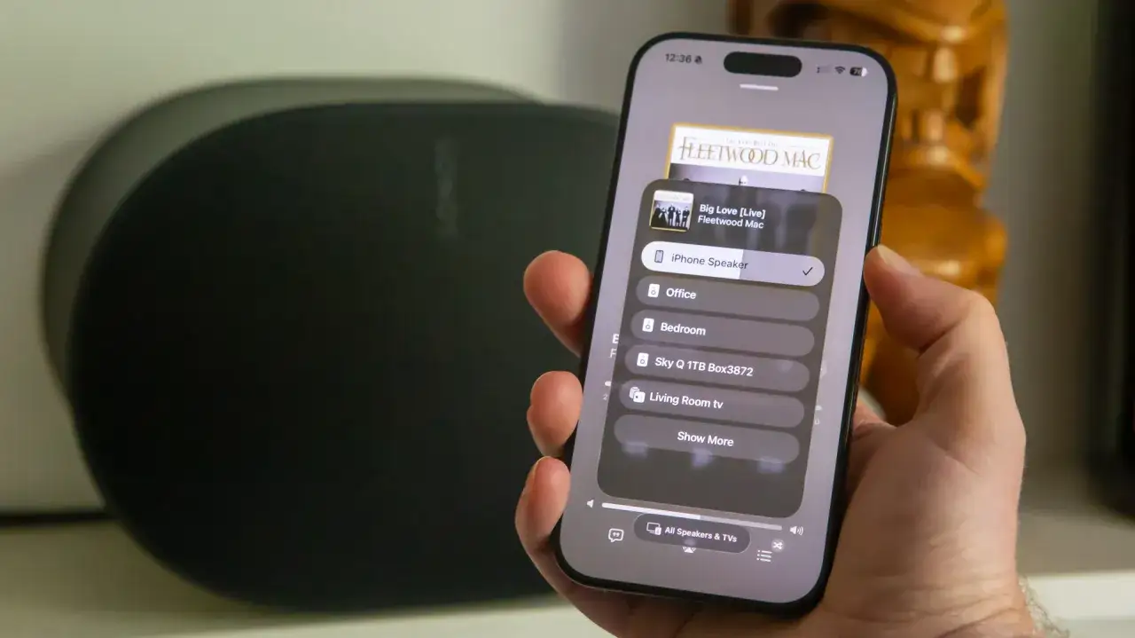 Dłoń trzyma smartfon z menu wyboru głośnika do **airplay**. Wyświetla się lista urządzeń, w tym