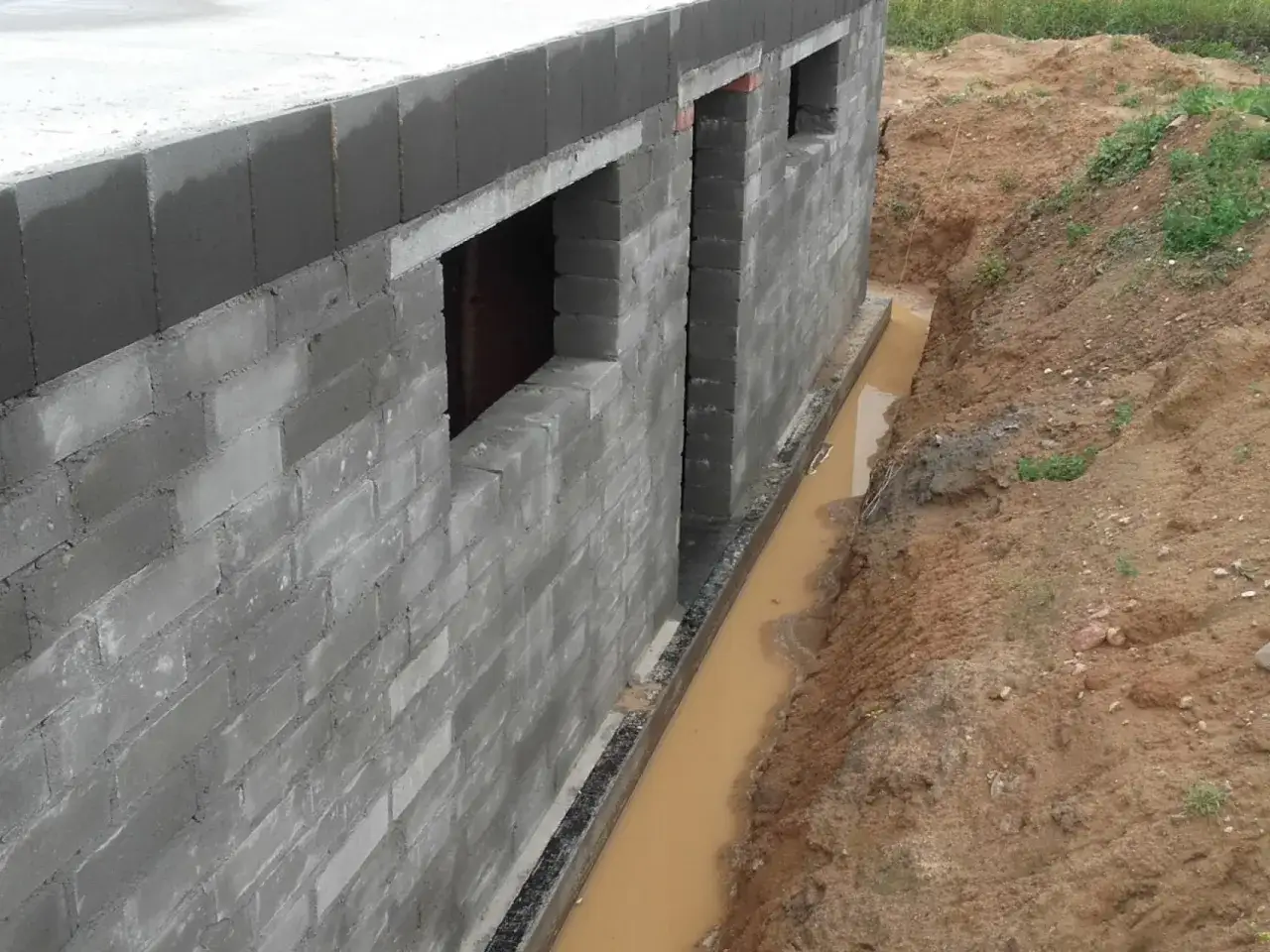 Budowa domu z bloczków betonowych, w wykopie widoczny jest błotnisty **drenaż** odprowadzający wodę.