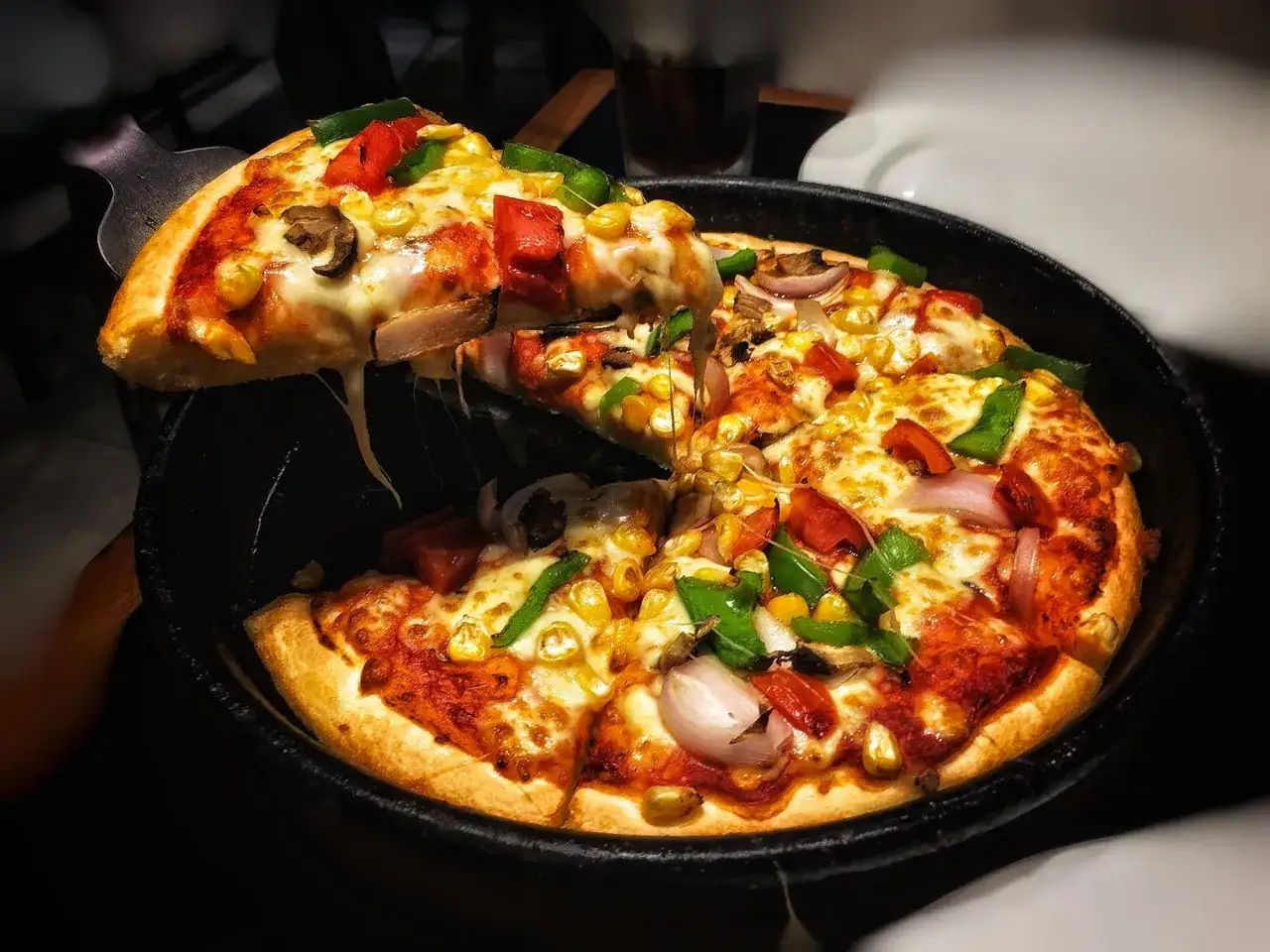 Una fetta di pizza in padella, filante e ricca di verdure, viene sollevata, invitando a gustare questo piatto delizioso.