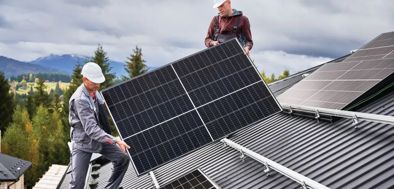 Zwei Handwerker installieren Solarmodule auf einem Dach, um die PV-Anlage zu erweitern. Im Hintergrund Berge und B&auml;ume.