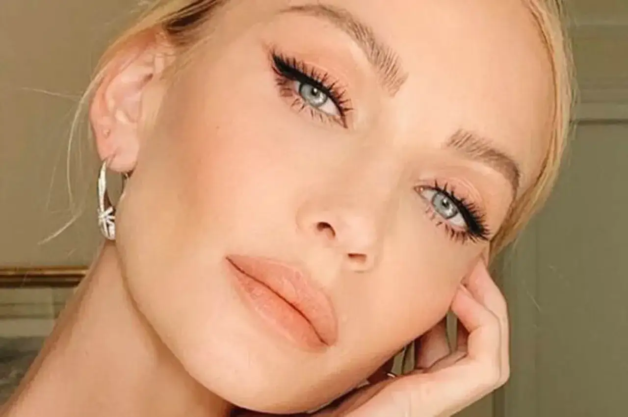 Primo piano di una donna con un trucco elegante: occhi azzurri con eyeliner e ciglia lunghe, labbra nude e orecchini a cerchio.