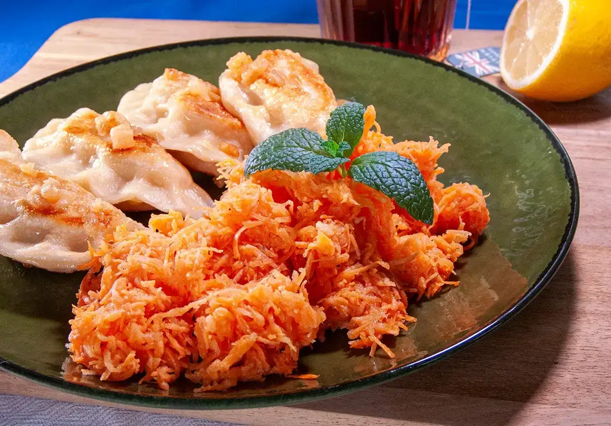 Na talerzu pierogi z rumianym sosem i świeża surówka z marchwi i pietruszki, udekorowana listkiem mięty.