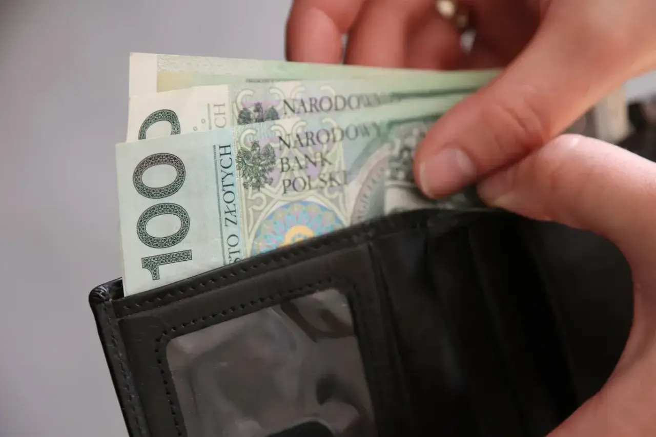 Ręce wkładają banknoty 100 zł do czarnego portfela. To może być dodatek za wysługę lat w budżet&oacute;wce.
