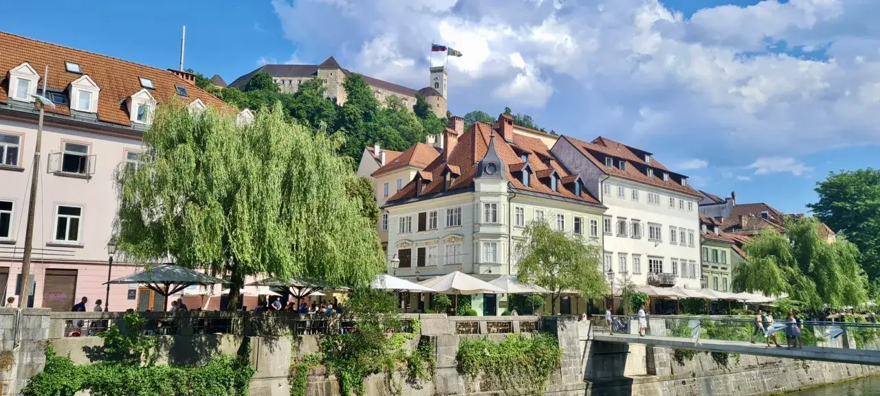Lublana atrakcje: Zamek na wzg&oacute;rzu, kolorowe kamienice nad rzeką, kawiarnie z ogr&oacute;dkami i zielone drzewa tworzą malowniczy widok.
