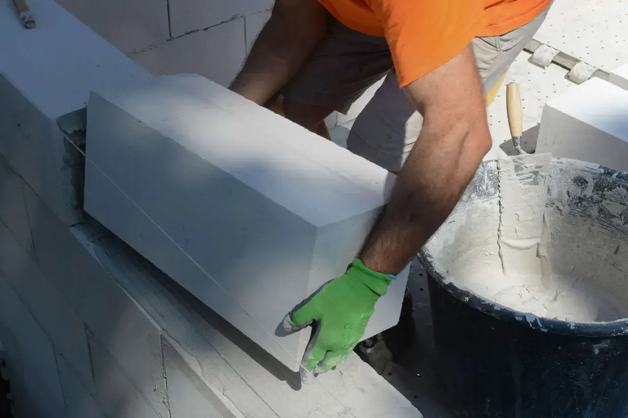 Mano con guanto verde posiziona un blocco di calcestruzzo aerato autoclavato su una parete in costruzione.