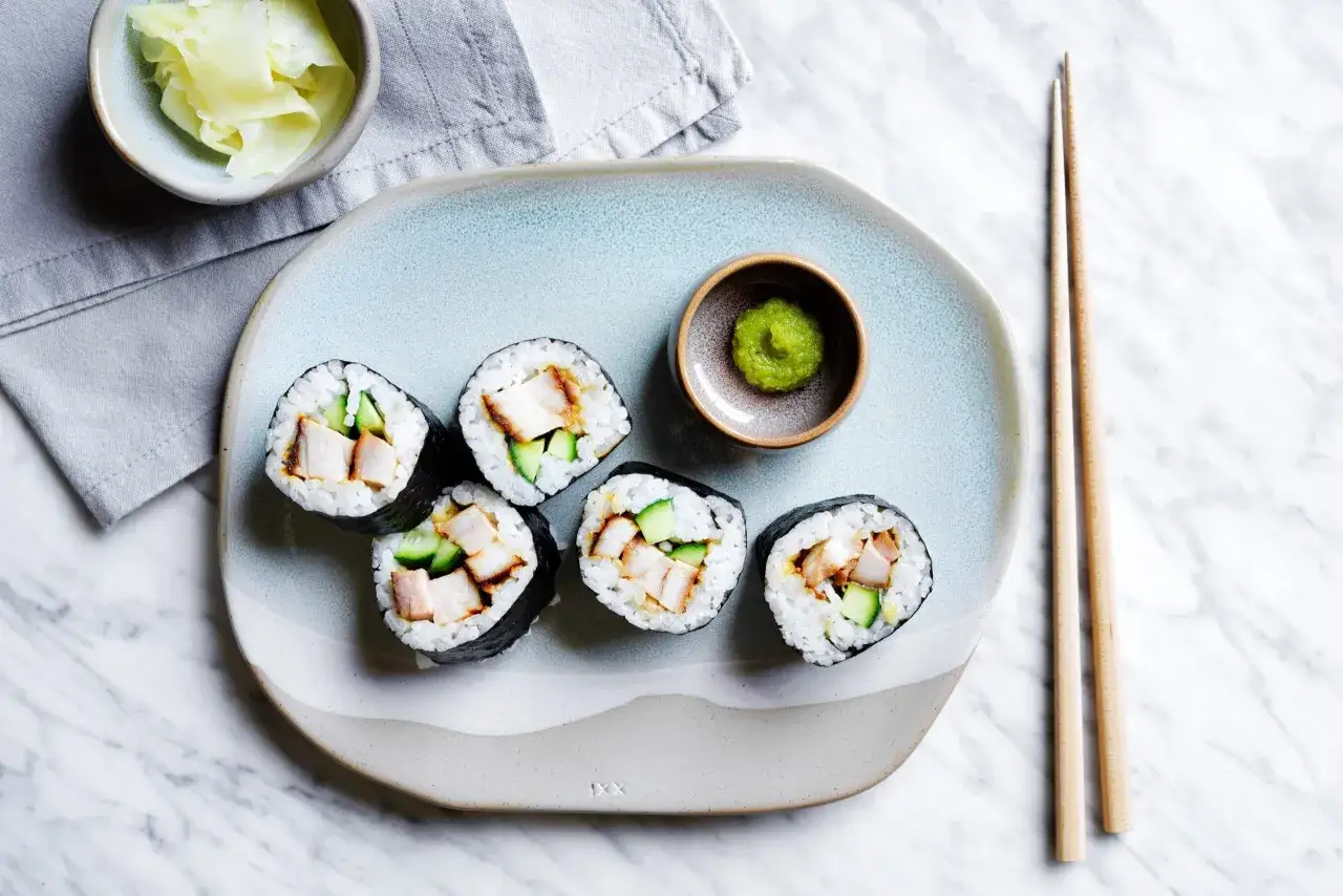 Pięć kawałków sushi z kurczakiem i ogórkiem na talerzu, obok miseczka z wasabi i marynowanym imbirem.