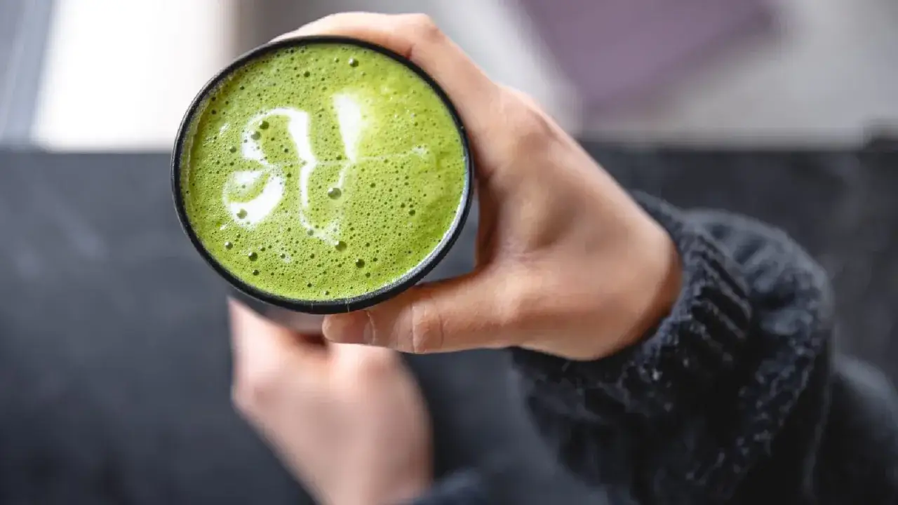 Grüner Matcha-Latte mit schaumiger Krone und einem Blattmuster. Eine Hand hält die Tasse, die für eine gesunde matcha schwangerschaft geeignet ist.