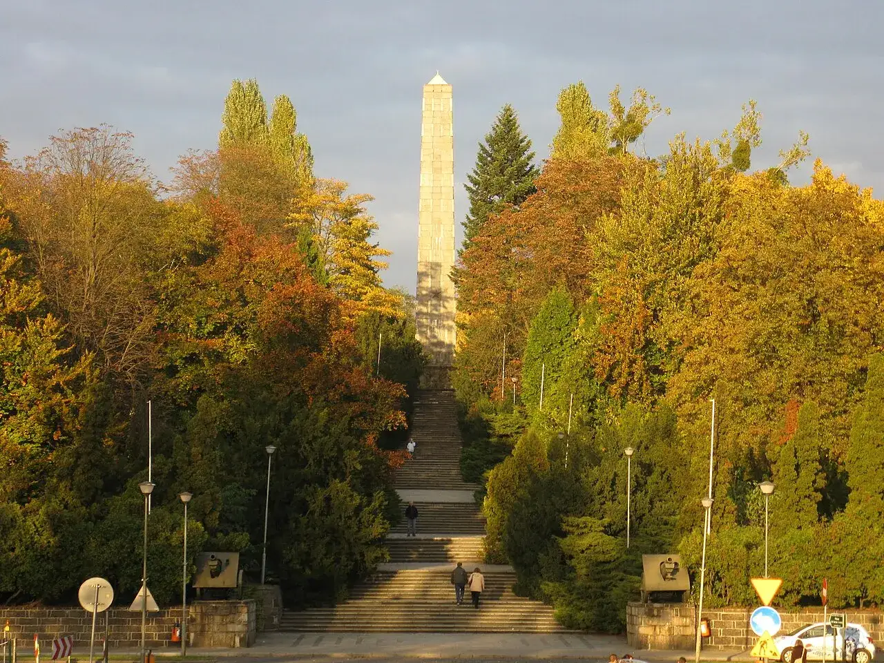 Jesienna panorama Poznania z monumentalnym obeliskiem i schodami. Wśród kolorowych drzew widać pomniki.