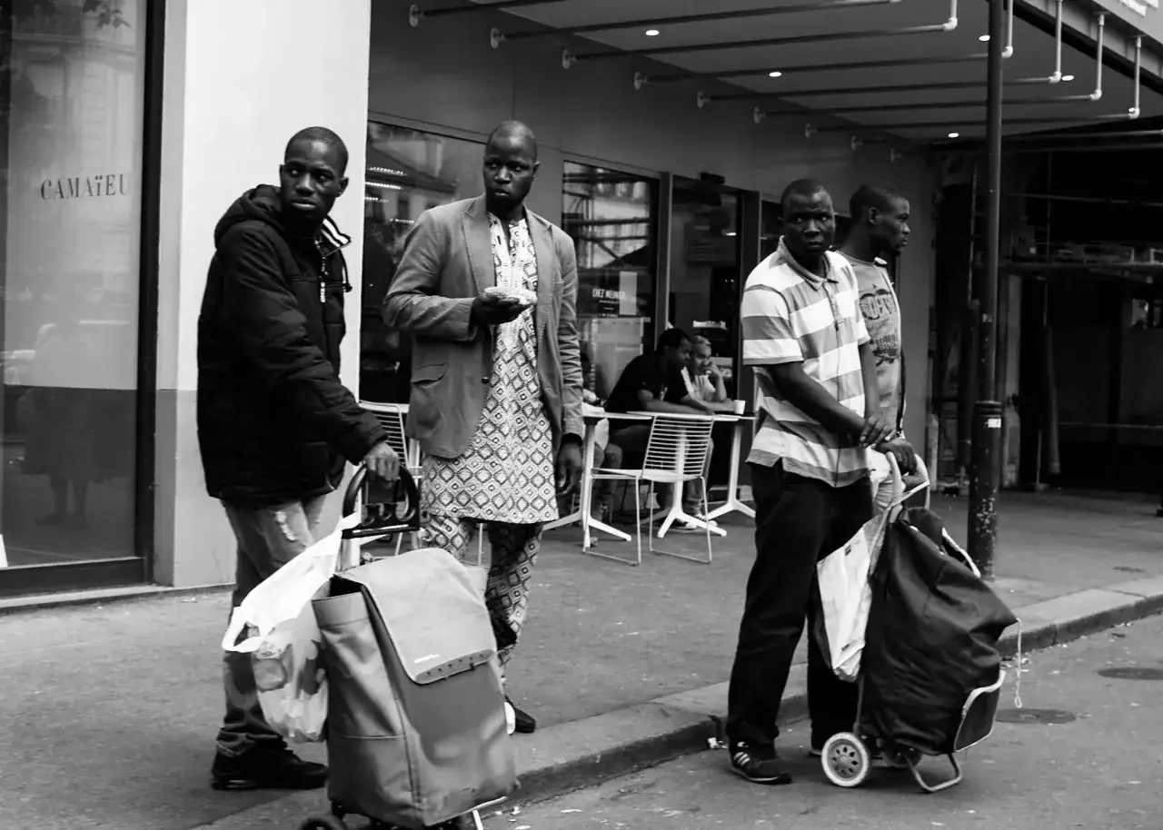Photographie de rue : quatre hommes, certains avec des chariots, attendent sur un trottoir devant un magasin.