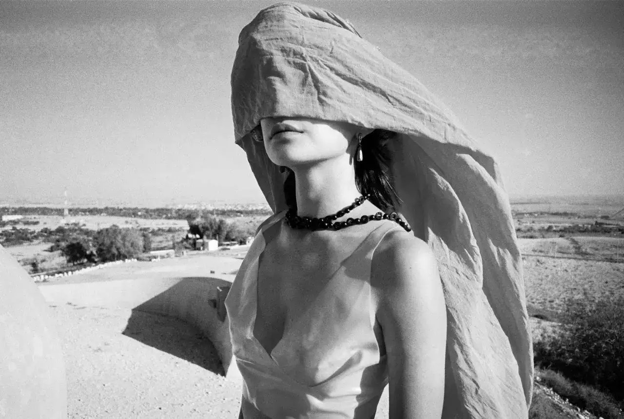 A woman with a light fabric covering her face, wearing a beaded necklace, stands against a hazy landscape. Shot on Ilford Pan 400.