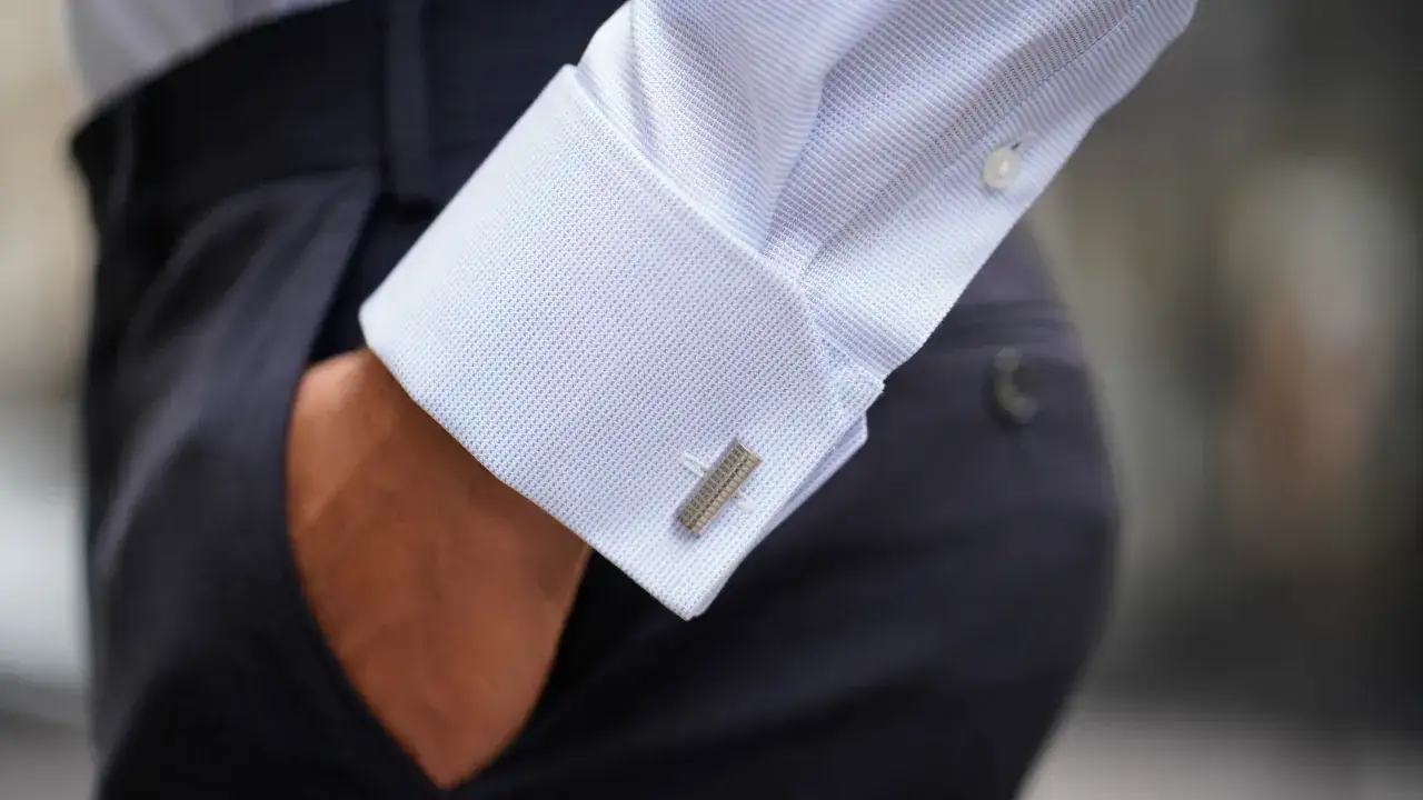 Détail d'une chemise manchette homme bleu clair avec boutons de manchette argentés, portée avec un pantalon bleu marine.