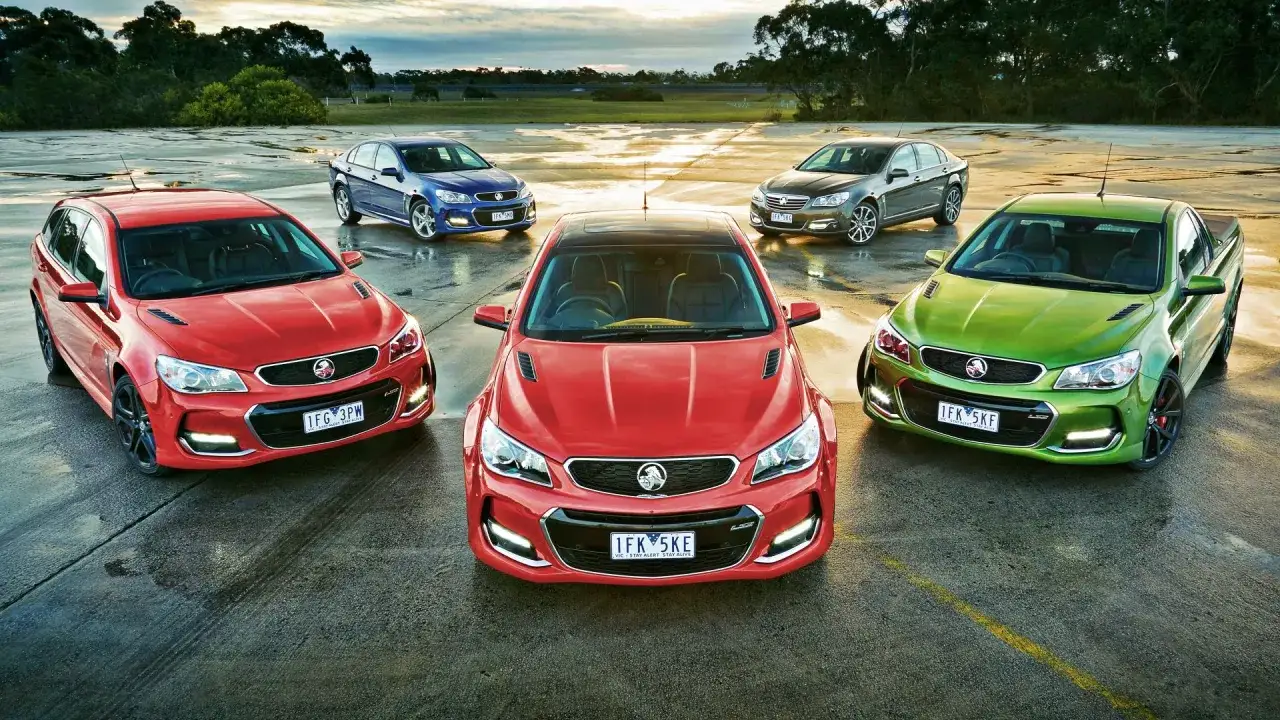 Kolekcja samochodów marki General Motors: czerwony sedan, zielony pickup, niebieski sedan, szary sedan i czerwony kombi na mokrej nawierzchni.