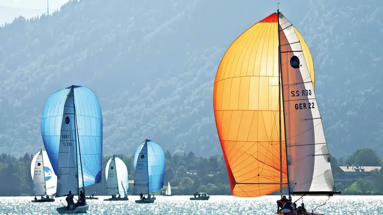 Segelboote mit bunten Spinnakern auf dem Wasser. Ein orangefarbenes Segel dominiert, während im Hintergrund blaue Segel und Berge zu sehen sind. Perfekt für einen Podcast über Segeln.