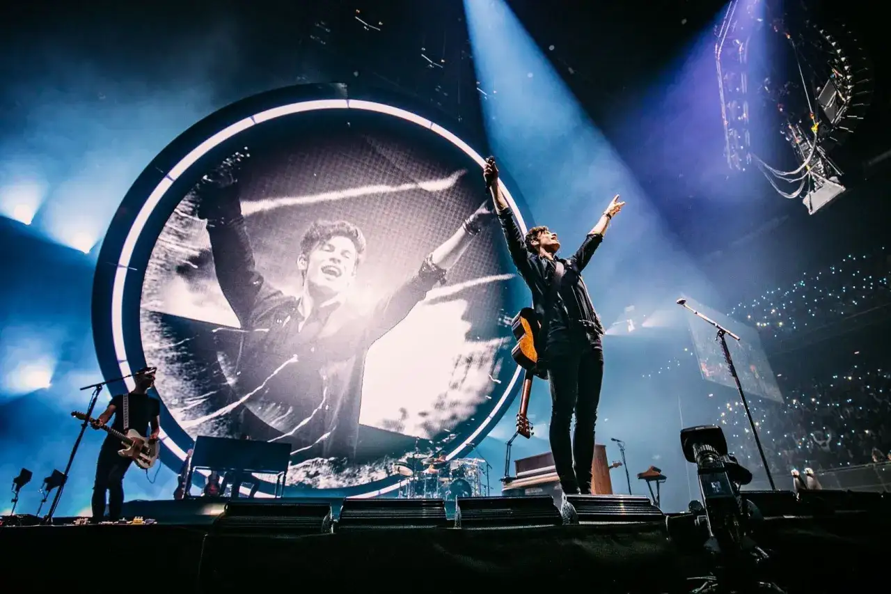 Shawn Mendes koncert, artysta z podniesionymi rękami na tle wielkiego ekranu z jego zdjęciem. Tłum świeci światłami.