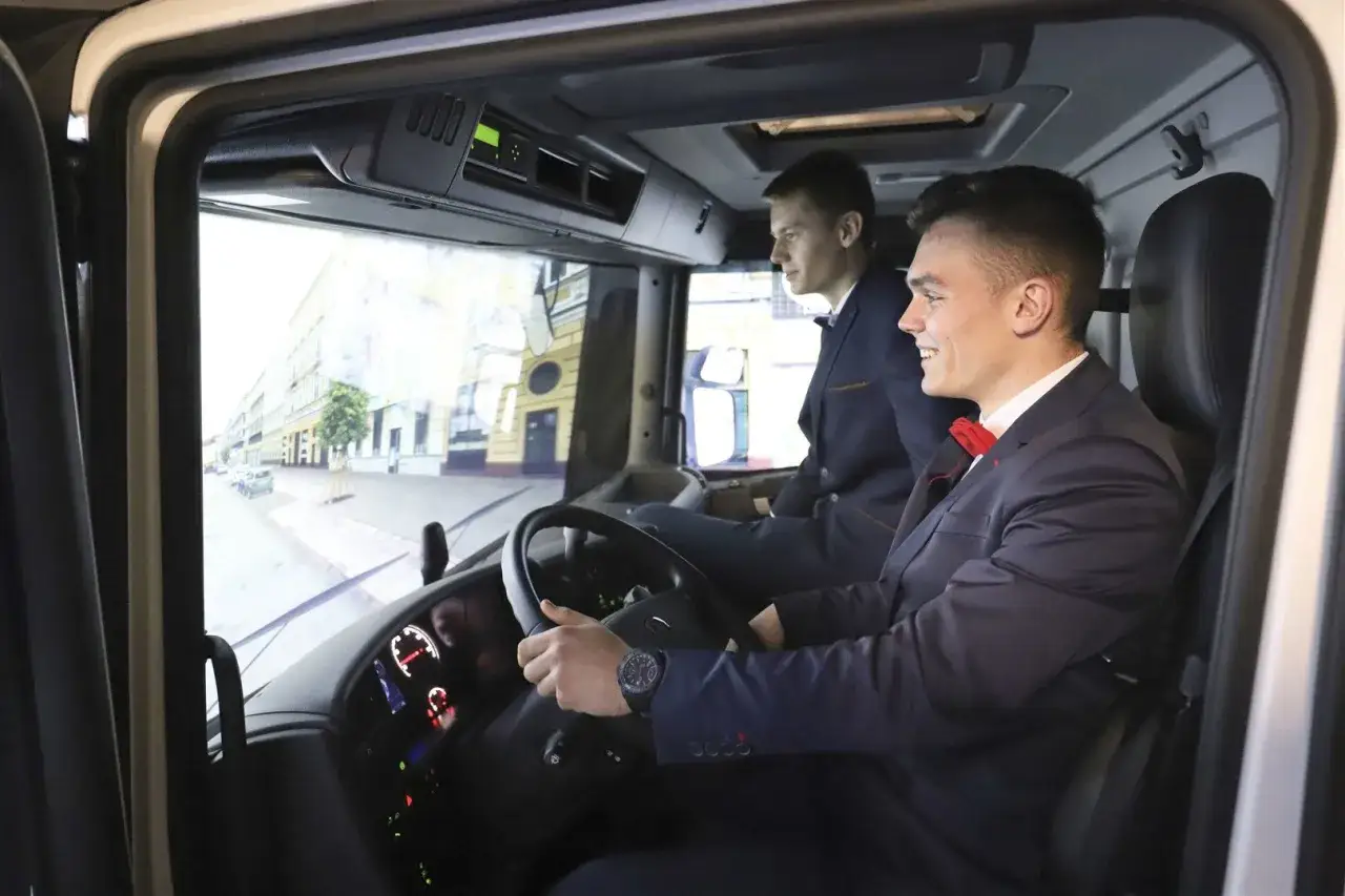 Dwóch młodych mężczyzn w garniturach siedzi w kabinie tira, ćwicząc jazdę. Zastanawiasz się, ile zarabiają kierowcy tira w Polsce?