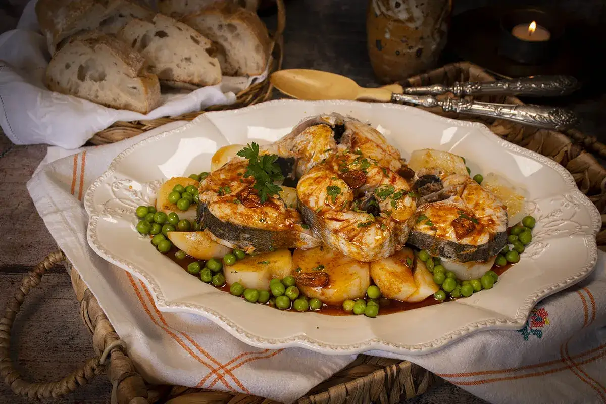 Merluza a la gallega servida con patatas y guisantes, decorada con perejil fresco. Pan rústico y una vela completan la escena.