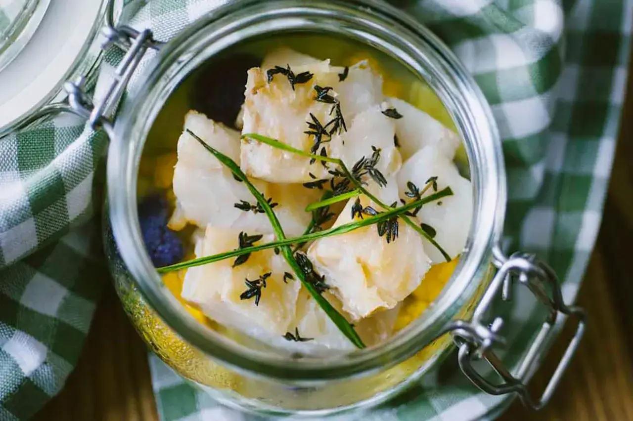 Pezzi di baccalà marinato in un barattolo di vetro, guarniti con erbe aromatiche. La differenza tra baccalà e stoccafisso è un tema culinario.