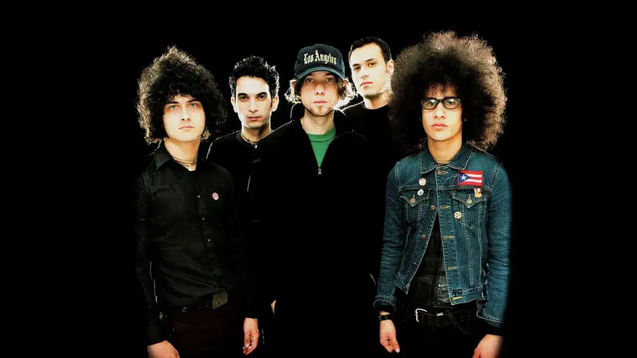 Cinq musiciens, dont deux avec des chevelures afro, posent pour une photo de groupe. L'un porte une casquette 