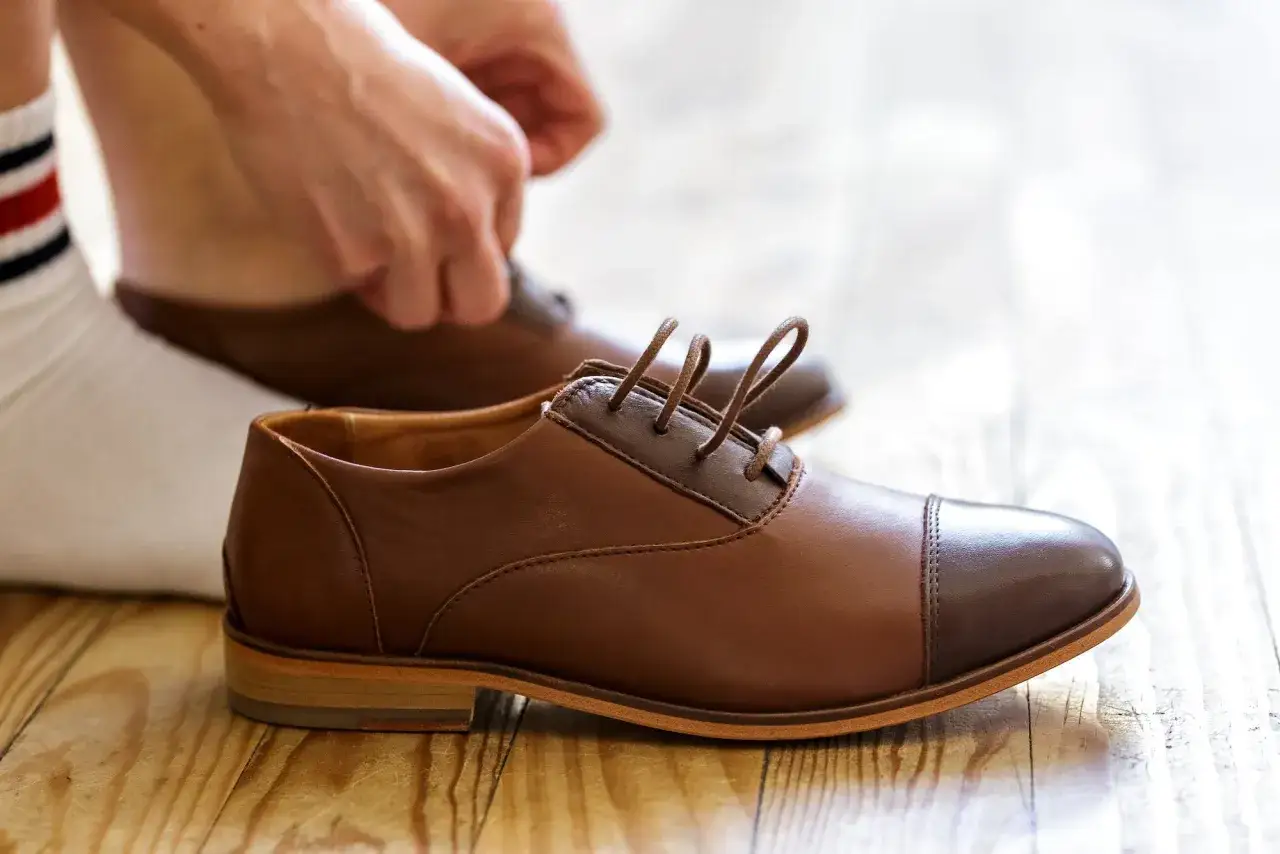 Mains ajustant les lacets de chaussettes derbies marron sur un parquet en bois.