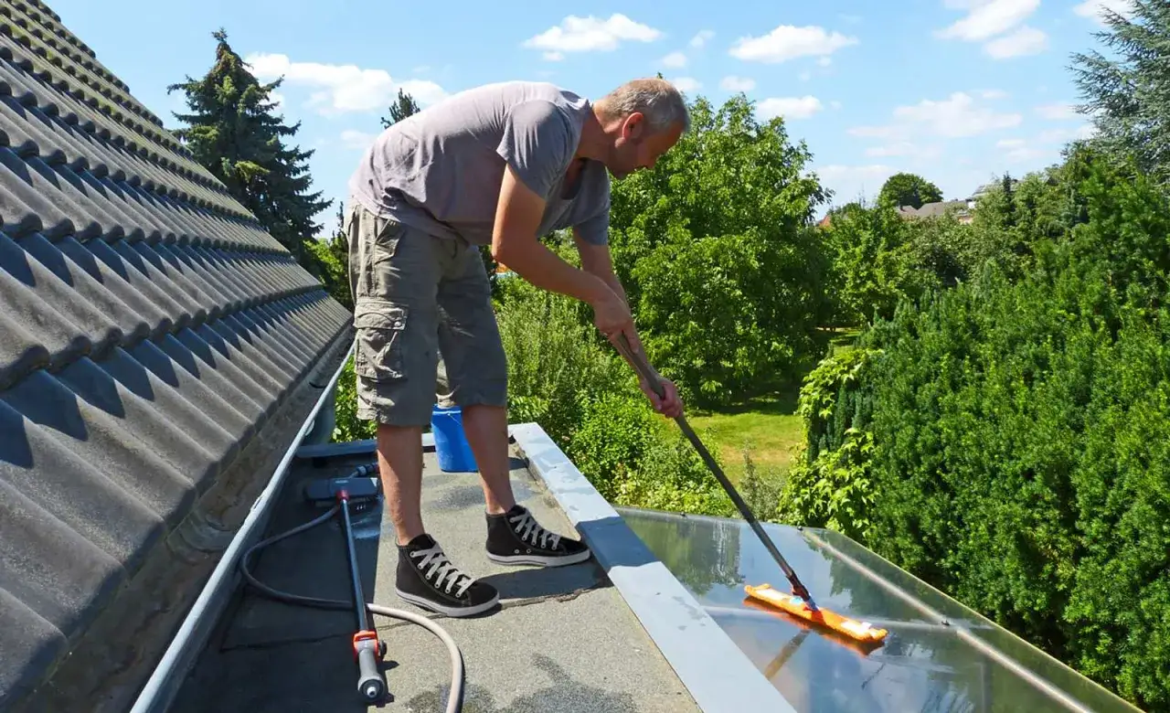 Mann reinigt mit einem Wischmopp eine Glasfläche auf dem Dach. Nebenan liegt die Dachpappe, die ebenfalls gereinigt werden muss.