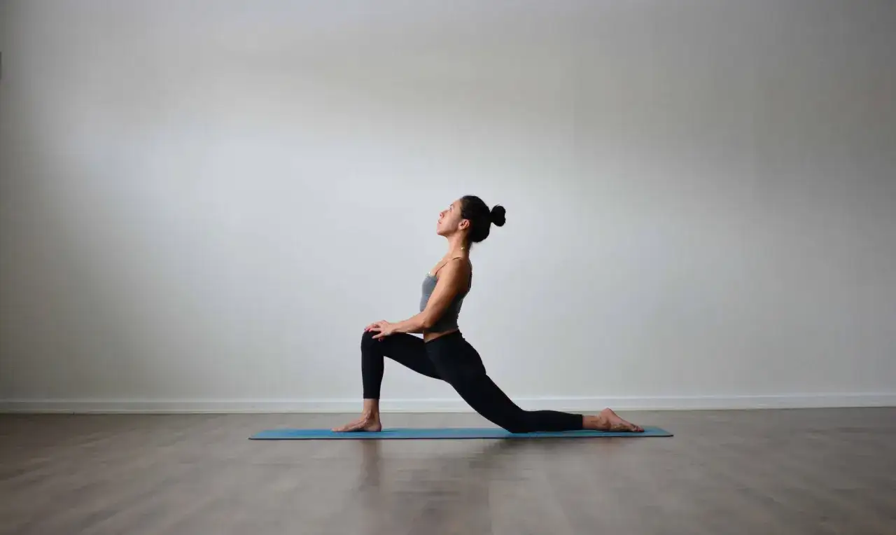 Mujer en postura de anjaneyasana yoga, con rodilla derecha flexionada y pie izquierdo extendido atrás.